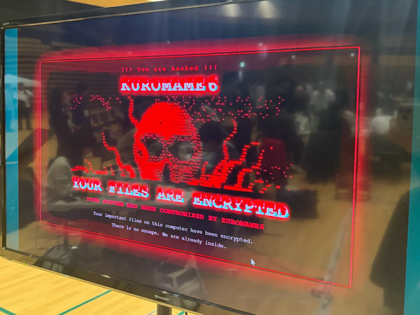 A monitor at the venue displaying Kuromame 6's "hacked" screen: a skull graphic alongside a message declaring the system's files encrypted.