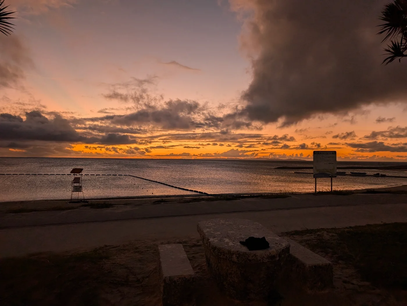 Sunset over the water at an Okinawan beach, a lifeguard chair and a notice board silhouetted against the orange sky.